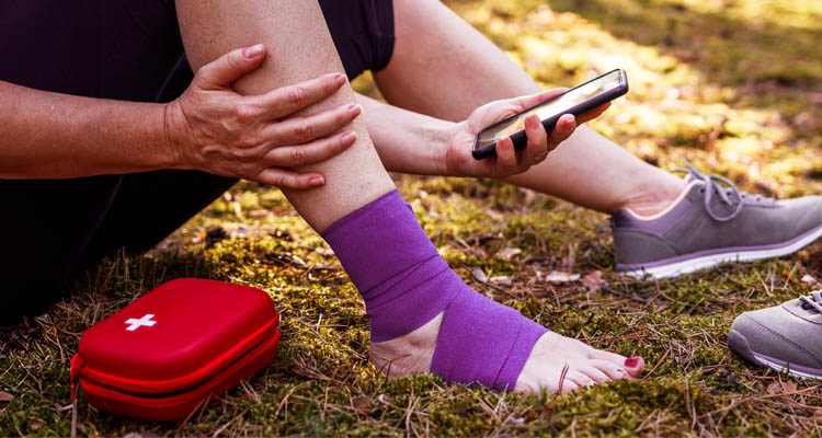 Eine Person sitzt im Freien, hat den Knöchel mit einer lila Bandage verbunden und hält ein Smartphone in der Hand, daneben liegt ein rotes Erste-Hilfe